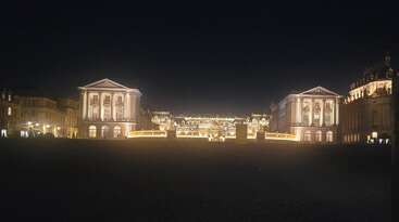 Cette image montre un palais illuminé la nuit, avec une architecture classique, de grandes façades, des lumières dorées, des bâtiments symétriquement alignés et un premier plan sombre et étendu.