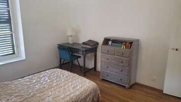 Cette chambre simple comprend un lit avec une couverture beige, un petit bureau et une chaise, une lampe, une commode avec des jouets et des planchers en bois.