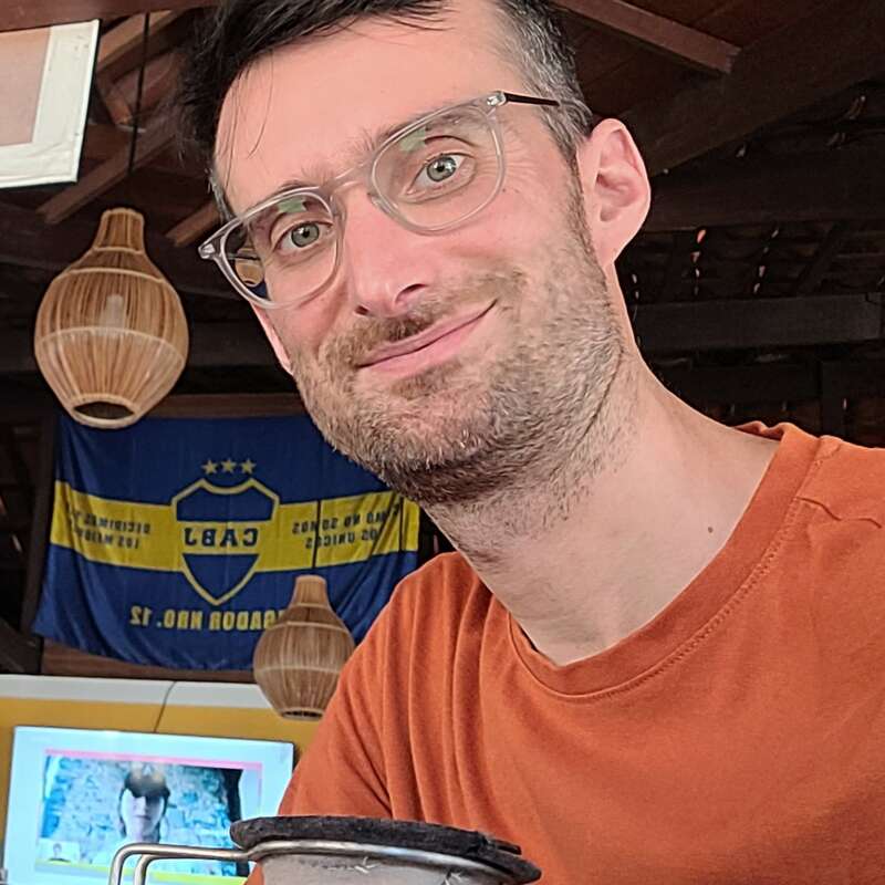 Un homme à lunettes et à la barbe taillée sourit à la caméra à l'intérieur. Derrière lui se trouvent des lampes suspendues, un drapeau bleu-jaune et un écran de télévision.