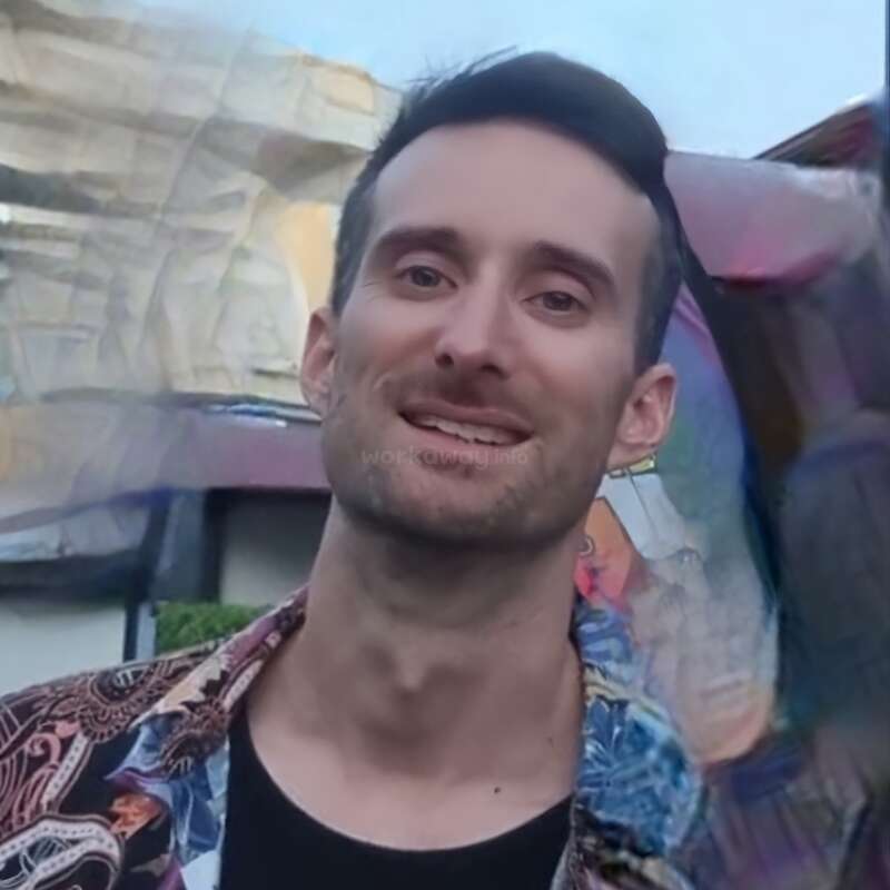 Un homme souriant aux cheveux bruns courts se tient à l'extérieur. Il porte une chemise colorée. L'arrière-plan présente des rochers et un effet artistique flou. Le ciel est bleu.