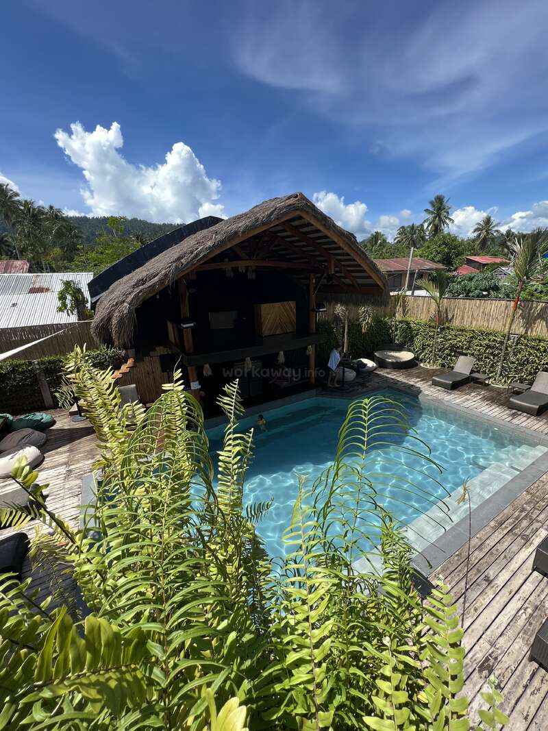 The image depicts a serene poolside scene with a small pool, a thatched-roof hut, and lounge chairs on a wooden deck, surrounded by lush greenery and a blue sky.