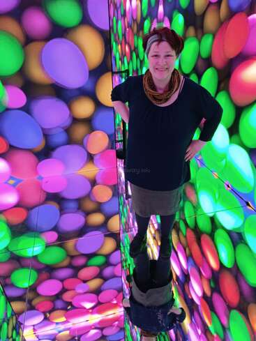 A smiling woman stands in a vibrant, colorful room with circular lights on the walls. Her reflection appears below her on a shiny mirrored floor.