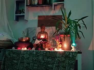 A serene altar with a Buddha statue, candlelight, plants, string lights, singing bowl, small flowers, and shelf with books, creating a peaceful and spiritual ambiance.