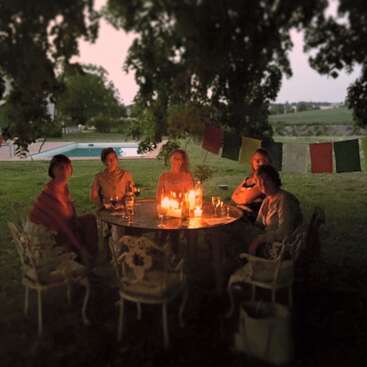 The image depicts a group of five people seated around a table, surrounded by candles, wine glasses, and a serene outdoor setting.