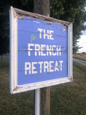 Une pancarte en bois bleu patiné porte l'inscription « THE FRENCH RETREAT » en lettres blanches. Elle est fixée sur un poteau, à l'extérieur, près d'une pelouse, d'arbres et d'une route de campagne.
