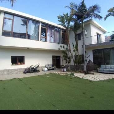 The image depicts a modern white house with a spacious yard, featuring a balcony, palm trees, and a patio area, set against a clear blue sky.