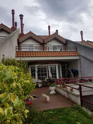 Ein gemütliches Stadthaus mit drei spitzen Dächern und Schornsteinen, einer Terrasse mit Stühlen, verstreutem Spielzeug, einem kleinen Hund, Pflanzen und einem bewölkten Himmel über uns. Die Wäsche trocknet.