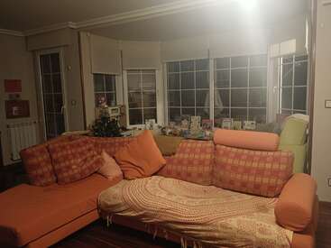 Ein gemütliches Wohnzimmer mit einem orange gemusterten Sofa, großen Fenstern, vielen Kissen, einer Decke und einer dekorierten Fläche hinter dem Sofa, auf der Karten und Fotos liegen.