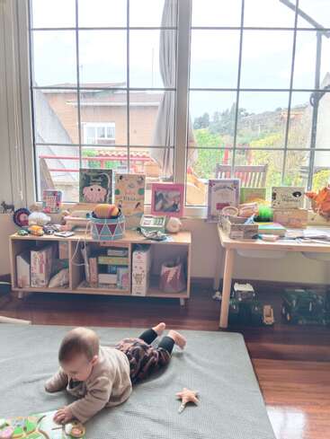 Ein Baby liegt auf einer Matte und spielt mit Spielzeug in einem gemütlichen, hellen Zimmer. Bücher und weiteres Spielzeug stehen in Regalen unter einem großen Fenster mit Aussicht.