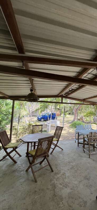 Eine überdachte Terrasse mit Holzmöbeln blickt auf einen Hof mit Palmen, einem blauen Auto und weiteren Sitzgelegenheiten im Freien. Die Umgebung wirkt entspannt und tropisch.