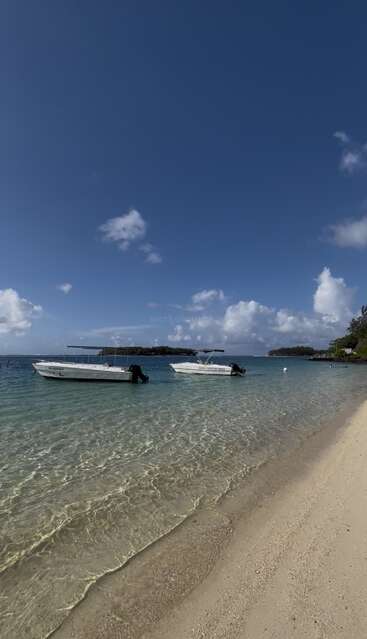 Klarer blauer Himmel, flauschige weiße Wolken, zwei Boote, die auf dem kristallklaren türkisfarbenen Meer treiben, sanfte Wellen, die den Sandstrand erreichen, ein friedliches tropisches Paradies, üppiges Grün in der Ferne.