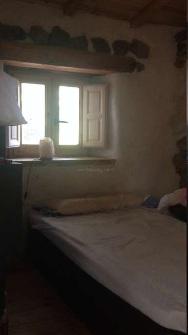 Das Bild zeigt ein schwach beleuchtetes Zimmer mit einem Bett, einem Fenster und einer Lampe, mit einem weißen Laken und Kissen und einer Steinwand im Hintergrund.