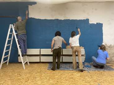 Cuatro personas están pintando de azul una pared blanca con textura. Una utiliza una escalera, dos usan rodillos de pie y una se arrodilla. El suelo está cubierto con láminas protectoras.