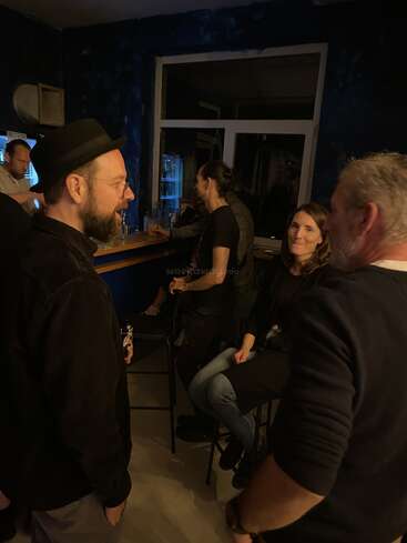 Un grupo de personas socializa en un bar poco iluminado. Algunos están de pie, otros sentados, conversando, con bebidas en la mano, creando un ambiente relajado y amistoso.