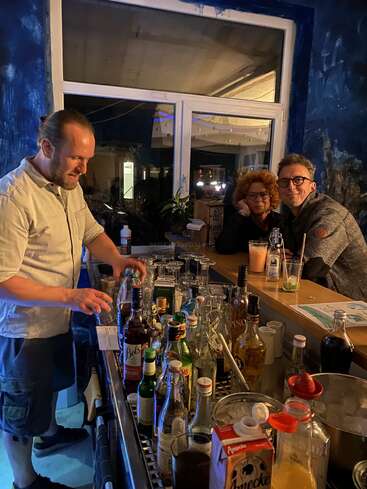 Un camarero prepara bebidas detrás de una barra desordenada con varias botellas, mientras dos personas se sientan juntas en la barra, sonriendo en un ambiente acogedor y poco iluminado.