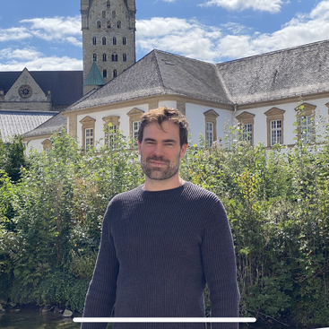 A man stands outdoors, smiling gently, in front of an old building with white walls, arched windows, and a tall stone tower under a blue sky.