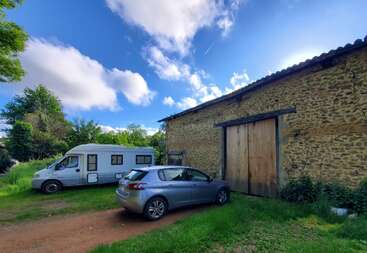 Un coche plateado y una autocaravana blanca están aparcados junto a un granero rústico de piedra, rodeados de vegetación bajo un cielo azul intenso con algunas nubes dispersas.