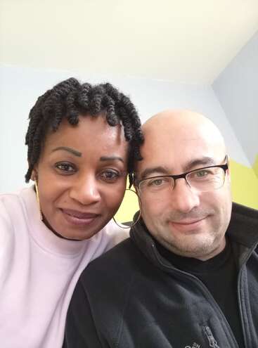 Una pareja sonriente posa muy junta para hacerse un selfi en el interior. La mujer tiene el pelo corto y rizado y lleva pendientes de aro dorados, mientras que el hombre lleva gafas.