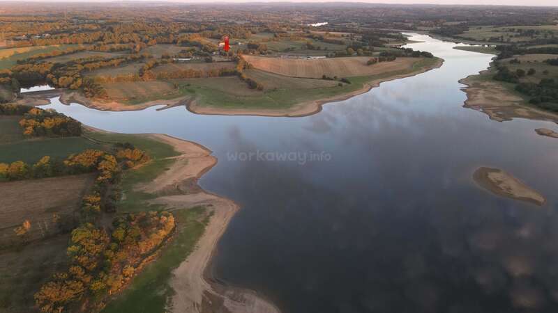 Una vista aérea de un río sinuoso o un lago rodeado de verdes campos, bosques y tierras de cultivo bajo la luz dorada del sol. Un punto rojo señala un lugar lejano.