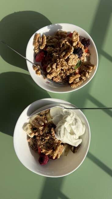 Auf einem grünen Tisch stehen zwei weiße Schalen, gefüllt mit Müsli, Walnüssen, Beeren und Joghurt, und werden von Sonnenlicht beschienen. In jeder Schale liegt ein Metalllöffel.