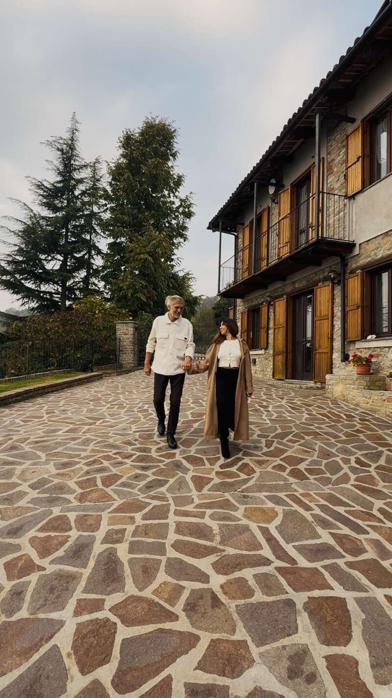 Ein Paar spaziert Hand in Hand über eine Steinterrasse vor einem charmanten Haus mit Holzfensterläden, umgeben von Bäumen, das Wärme und Ruhe ausstrahlt.