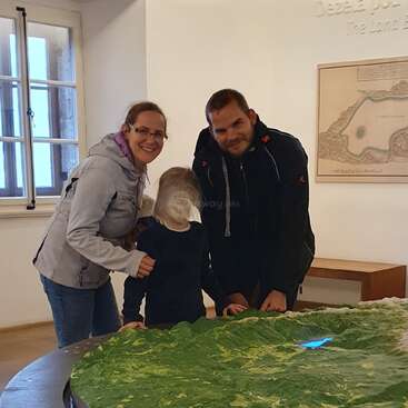Eine lächelnde dreiköpfige Familie posiert drinnen vor einer großen grünen 3D-Karte. Die Erwachsenen stehen dicht beieinander, während das Kind zwischen ihnen steht und die Karte betrachtet.