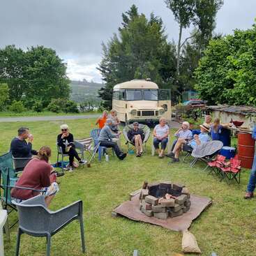 A imagem mostra um grupo de pessoas reunidas em torno de uma fogueira em uma área gramada, com um ônibus e árvores visíveis ao fundo em um dia nublado.