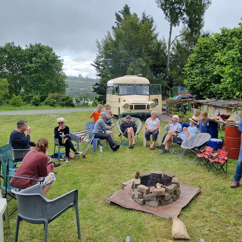 L'image représente un groupe de personnes réunies autour d'un feu dans un espace herbeux, avec un bus et des arbres visibles à l'arrière-plan par temps couvert.