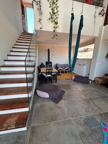 Ein moderner Wohnraum mit gefliestem Boden, Holztreppe mit Glasgeländer, hängenden Grünpflanzen, türkisfarbener Luftseide, gemütlicher Sitzecke und Bodenkissen. Helle Atmosphäre.