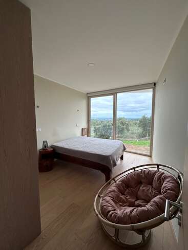 This image shows a minimalist bedroom with a bed, wooden floor, a cozy papasan chair, and large windows offering a scenic view of greenery and sky.
