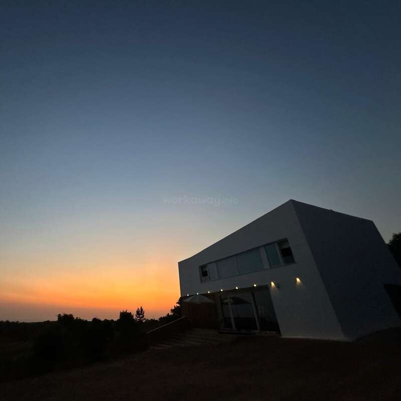 Un moderno edificio blanco se asienta en un ángulo bajo un cielo crepuscular, con cálidas luces iluminando su lateral. La puesta de sol crea un horizonte tranquilo y colorido.
