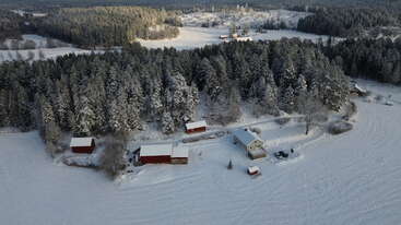 Vue aérienne d'un paysage rural enneigé avec plusieurs bâtiments rouges et blancs, entouré d'une forêt dense. Traces dans la neige, paysage hivernal serein, village lointain.