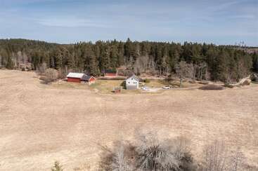 Vue aérienne d'une ferme rurale avec une maison blanche, des granges rouges, une forêt environnante et des champs ouverts sous un ciel dégagé, respirant la tranquillité de la campagne.