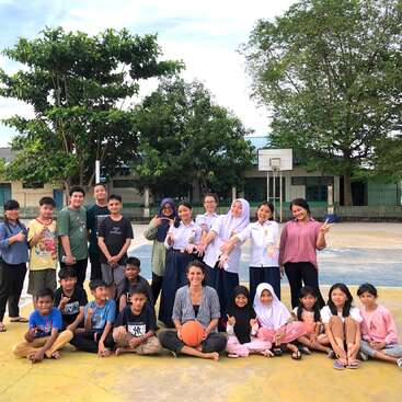 A group of students and teachers pose on an outdoor basketball court, smiling cheerfully. Some are sitting, some standing, with trees and a school in the background.