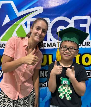 A smiling woman and a boy, wearing a green graduation cap, stand together in front of a colorful backdrop, giving enthusiastic thumbs up to celebrate success.