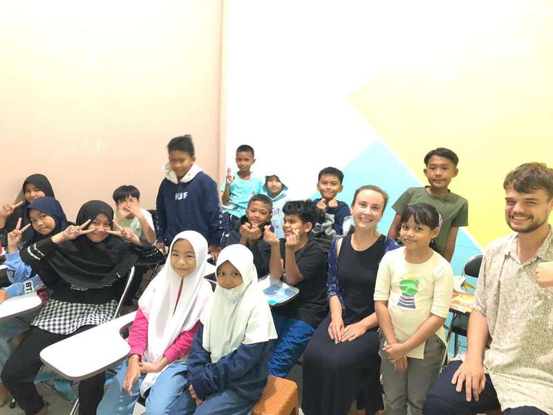 The image depicts a group of children and an adult posing for a photo in a classroom setting, with a diverse range of clothing and facial expressions.