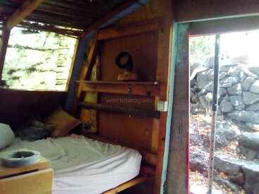 The image depicts a cozy wooden cabin with a bed, window, and door, situated in a natural setting with rocks and trees visible outside the open door.