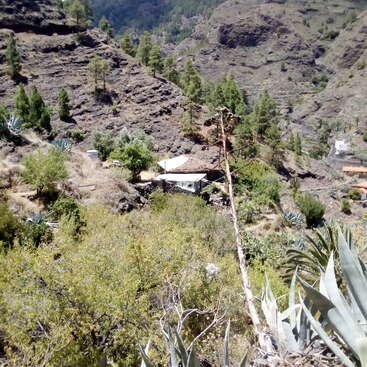 The image depicts a serene mountainous landscape with a white house nestled among trees and bushes, surrounded by rugged terrain and a distant valley.