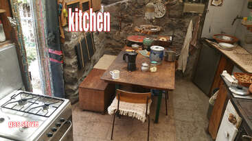 The image depicts a kitchen with a stove, sink, and cabinets, featuring a table and chairs in the background, set against a neutral-colored wall. The overall atmosphere is clean and organized.