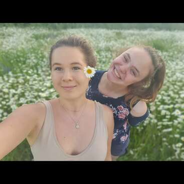 Two young women taking a selfie in a field of daisies, smiling happily. One has a daisy in her hair, both enjoying a summer day outdoors.