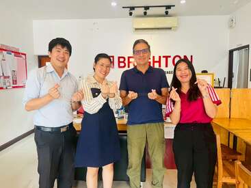 Four people are standing indoors, smiling, and making finger heart gestures. Behind them is a sign that says "BRIGHTON." The atmosphere is cheerful and welcoming.
