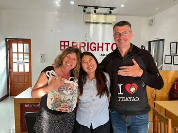 Three smiling people pose inside a language center. The woman on the left holds a cat-themed bag, the man on the right wears an “I ♥ Phayao” shirt.