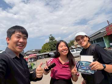 Three friends smile brightly outside on a sunny day, enjoying each other's company. Two give thumbs up, one holds a phone. Buildings and trees are behind them.