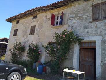 An old stone house with wooden shutters and a tiled roof. Pink roses climb the walls. A black car and outdoor table sit nearby under blue sky.