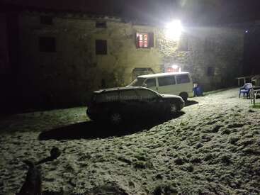 At night, two vehicles are parked on a snowy lawn in front of an old stone building, illuminated by a bright outdoor light. Blue chairs nearby.