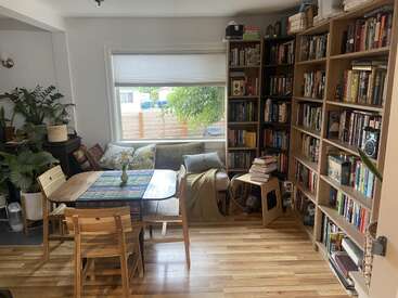 A cozy room with wooden floors, bookshelves filled with books, a dining table with chairs, a couch by the window, and several houseplants.