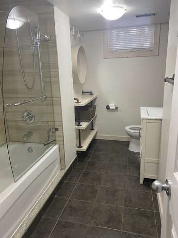 This bathroom features a bathtub with glass shower, dark tiled floor, modern sink with shelves, toilet, round mirror, window with blinds, and bright lighting.