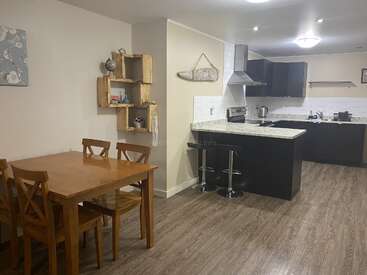 This image shows a modern kitchen and dining area with wooden floors, a wooden dining table, open shelving, bar stools, dark cabinets, and neutral decor.