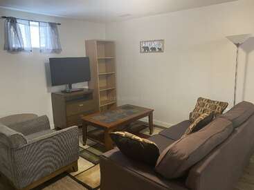 A cozy living room with a gray couch, armchair, wooden coffee table, TV, bookshelf, floor lamp, small window with curtains, and neutral-colored decor.