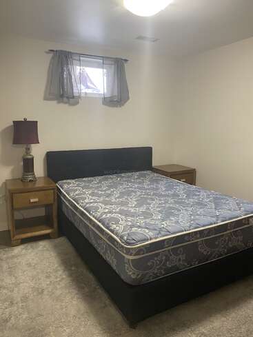 This is a simple bedroom with a bed, mattress, two wooden nightstands, a lamp, grey curtains, a small window, light walls, and carpeted flooring.